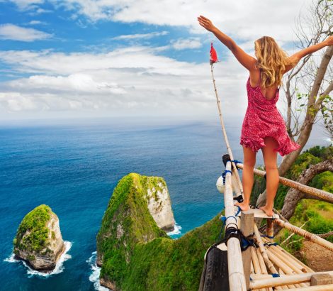 Family vacation lifestyle. Happy woman with raised in air hand stand at viewpoint. Look at Kelingking beach under high cliff. Travel destination in Bali. Popular place to visit on Nusa Penida island.